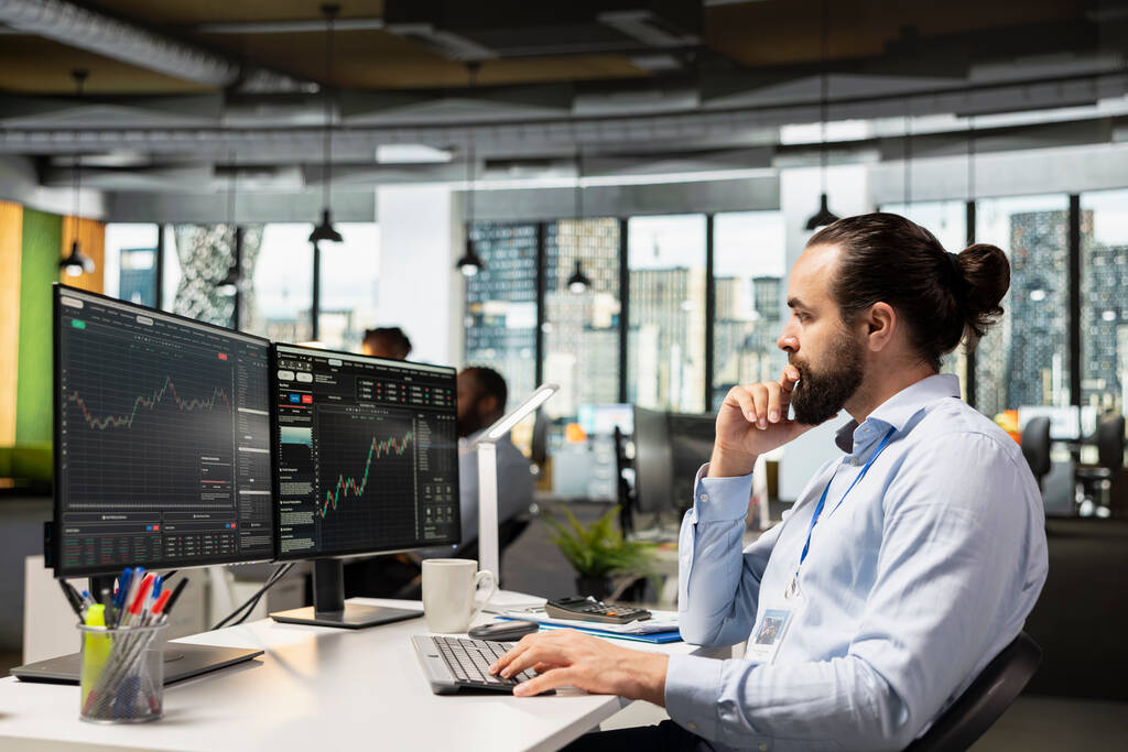Analista de TI em escritório moderno monitorando gráficos de conformidade e uso de licenças através de um sistema de Software Asset Management em dois monitores.