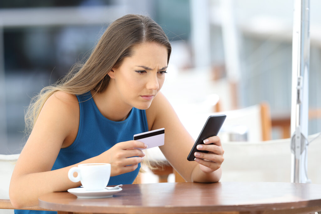 phishing o que e golpe cartao celular.jpg Mulher desconfiada analisando celular e cartão bancário em exemplo de phishing